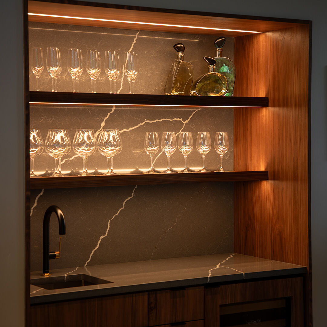 A wet bar complete with marble countertop, sink, and glasses on shelves.